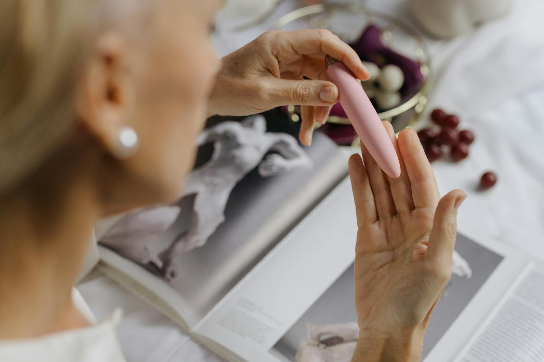 Fotografia de uma mulher a segurar um vibrador rosa, com livros de arte ao fundo, enfatizando temas íntimos e pessoais.