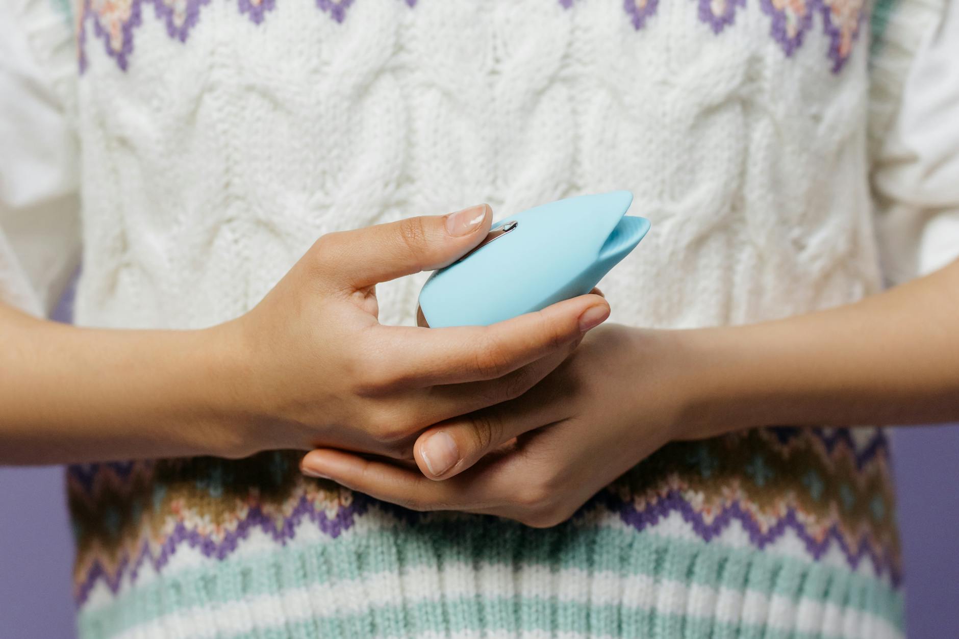 Close-up of hands holding a blue personal massager against a knitted sweater, representing comfort and self-care for pleasure recovery.